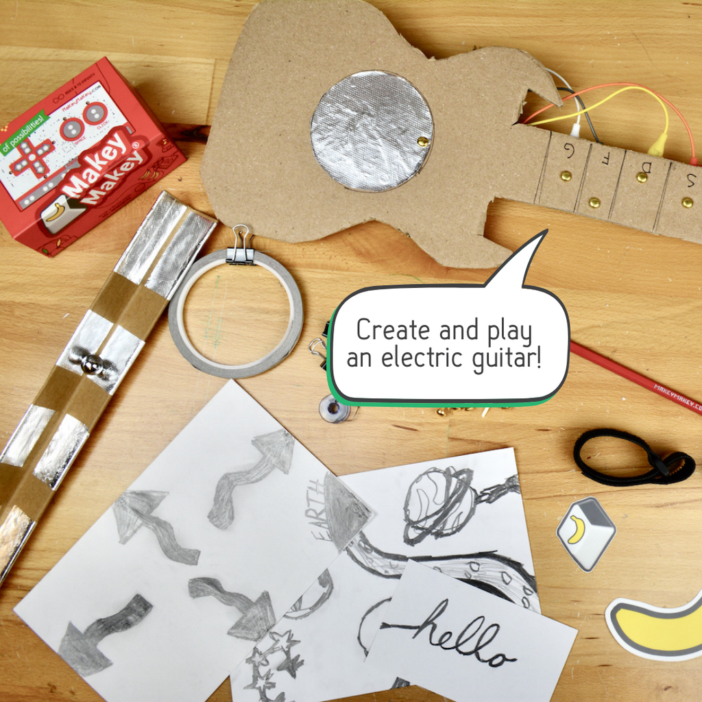 Handmade cardboard guitar with brass buttons and a callout bubble that says "Create and play an electric guitar" on a table.  Scattered on the desktop are more Craft + Code supplies and drawing projects on the table.